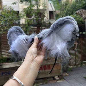 May include: A pair of fluffy, grey and white faux fur animal ears attached to a thin black headband. The ears feature long, soft fibers and a silver-toned metal chain accessory hanging from the right side, which includes a small cross charm.