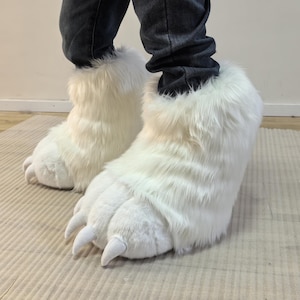 May include: White faux fur bear paw slippers with large claws. The slippers are designed to cover the feet and ankles, with a fluffy texture and a realistic claw design. These novelty slippers are a fun and unique accessory.