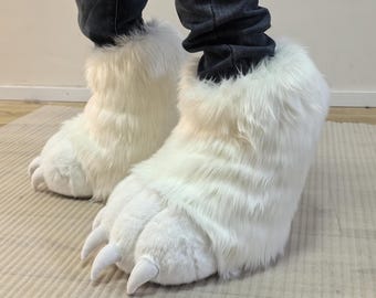 May include: White faux fur bear paw slippers with large claws. The slippers are designed to cover the feet and ankles, with a fluffy texture and a realistic claw design. These novelty slippers are a fun and unique accessory.