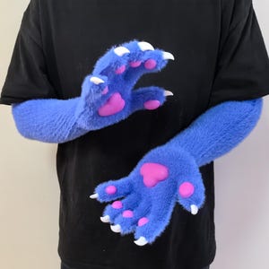Puede incluir: Un par de guantes de pata azules y peludos con almohadillas rosas y garras blancas. Los guantes están diseñados para parecerse a las patas de los animales, con una manga larga.