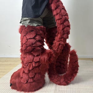 May include: A pair of deep red faux fur boots featuring a layered, scale-like texture and a matching long, thick, curved faux fur tail attachment. The boots have a bulky, rounded shape with small black claw-like details at the front toes.