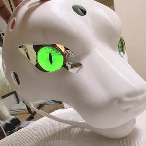 May include: A white fursuit head with large, glowing green eyes. The head features a smooth, rounded design with open eye sockets and small holes. The eyes are a bright green, providing a vivid contrast against the white surface.