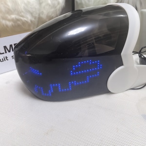 May include: A black and white futuristic helmet with a blue LED display showing a pixelated face. The helmet has a visor and ear pieces. The text "LM" is partially visible on a box in the background.