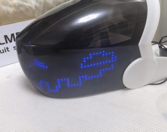 Puede incluir: Un casco futurista en blanco y negro con una pantalla LED azul que muestra una cara pixelada. El casco tiene una visera y auriculares. El texto "LM" es parcialmente visible en una caja en el fondo.