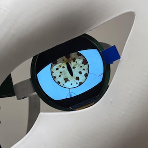 Op de afbeelding: Een close-up van een robotisch oog met een scherm dat een reptieleniris weergeeft. Het oog is geplaatst in een witte behuizing met een uitsparing. De iris is bruin met een zwarte pupil, tegen een lichtblauwe achtergrond.