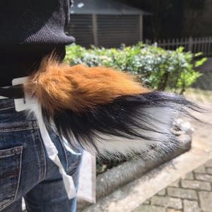 Puede incluir: Un accesorio de cola de animal de piel sintética con un patrón tricolor de marrón, negro y blanco. La cola de felpa está unida a un mecanismo de clip de plástico blanco diseñado para una fijación segura a la ropa, mostrando fibras sintéticas largas y suaves.