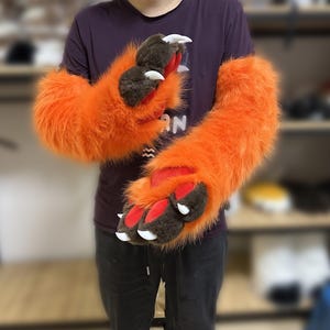 May include: Orange furry paw gloves with brown paw pads and white claws. The gloves have a red interior and are designed to resemble animal paws. The gloves are made of a soft, plush material.