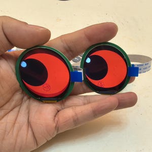 Puede incluir: Dos componentes electrónicos redondos, con forma de ojo, rojos y negros, con un borde verde y un acento azul. Cada componente tiene un diseño en forma de media luna y está conectado a un cable plano. Sostenidos en una mano.