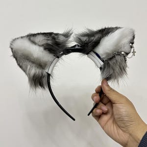 Op de afbeelding: Een zwarte haarband met twee imitatiebont dierenoren in wit-, grijs- en zwarttinten. Het rechteroor is versierd met een zilverkleurige metalen ketting en een kleine kruishanger, wat zorgt voor een gelaagd en gestructureerd uiterlijk.