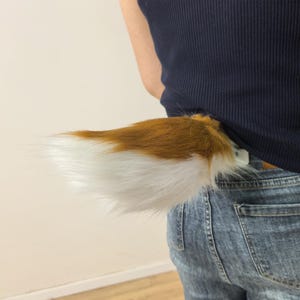 Peut inclure: Un accessoire de queue d'animal en fausse fourrure composé de fibres synthétiques à poils longs dans des tons marron et blanc. La queue est fixée à un débardeur côtelé bleu foncé et à un jean bleu, présentant un dégradé de couleurs réaliste.