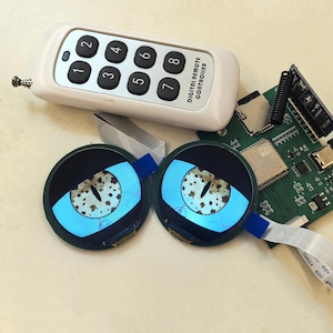 以下が含まれることがあります： 「DIGITAL REMOTE CONTROLLER」と書かれた白いリモコンが付いた、分解された電子機器。2つの円形スクリーンには、目の様な模様が表示されています。回路基板とリボンケーブルもあり、技術系またはDIYプロジェクトを示唆しています。