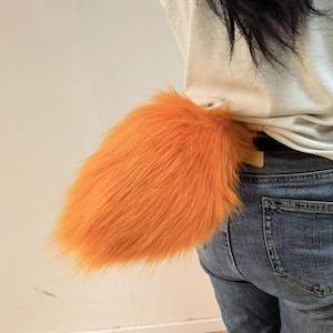 Puede incluir: Una cola de piel sintética de color naranja brillante y esponjosa sujeta a una camisa clara y unos vaqueros azules. La cola es gruesa, texturizada y presenta fibras sintéticas largas y suaves que crean una forma redondeada y voluminosa.