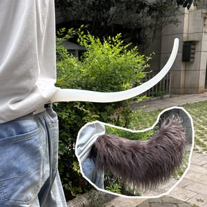 May include: A long, smooth, white, curved tail attachment worn at the waist, shown alongside an inset image of a long, shaggy, dark grey faux fur tail. The white tail is rigid and tapered, while the grey tail features dense, textured fibres.