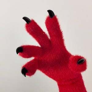 Op de afbeelding: Een rode, harige hand met zwarte klauwen. De hand is uitgestrekt met de vingers gespreid.