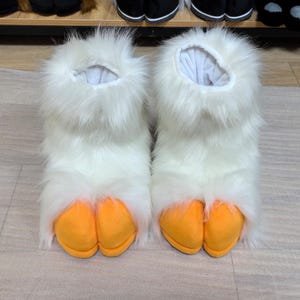 May include: A pair of white fluffy slippers with orange webbed feet. The slippers are designed to resemble duck feet.