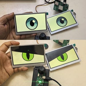 Op de afbeelding: Twee rechthoekige LCD-schermen verbonden met een printplaat en een joystickcontroller. De schermen tonen geanimeerde afbeeldingen van ogen, met een blauwe iris in de bovenste afbeelding en een groen katachtig oog met zwart ooglid onderaan.