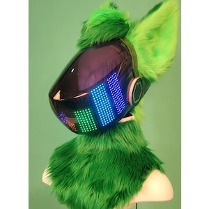 May include: Green furry head with a black helmet featuring a colourful LED display. The helmet has a black visor and a round, black earpiece on the right side.