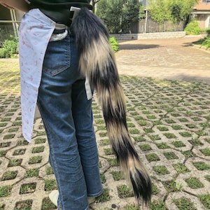 May include: A long, fluffy faux fur tail featuring a blend of black, grey, and cream-colored strands. The tail is attached to a black belt worn over blue denim jeans and a white fabric accessory with a subtle, light-colored pattern.