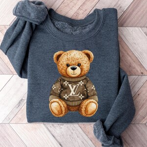 May include: A dark gray sweatshirt featuring a watercolor illustration of a teddy bear wearing a brown sweater with a white and brown logo. The bear has a friendly expression.