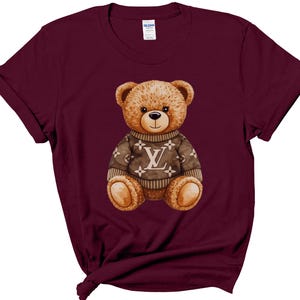 May include: A maroon t-shirt featuring a watercolor illustration of a teddy bear wearing a brown sweater with a white "LV" logo. The teddy bear has brown fur, a black nose, and is centered on the shirt.