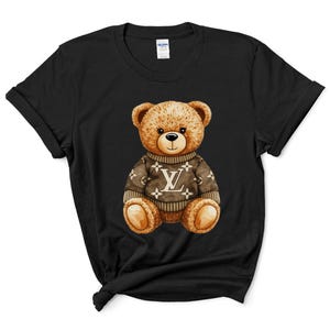 May include: Black t-shirt with a watercolour illustration of a teddy bear wearing a brown sweater. The sweater features a white logo design. The bear has brown fur and a black nose, centered on the shirt.