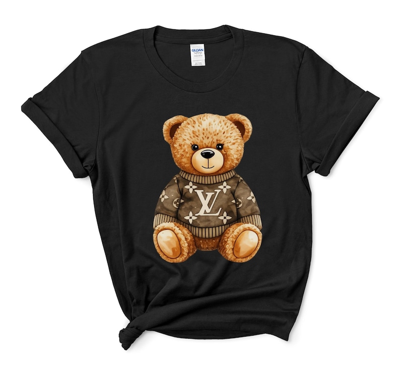 May include: Black t-shirt with a watercolour illustration of a teddy bear wearing a brown sweater. The sweater features a white logo design. The bear has brown fur and a black nose, centered on the shirt.