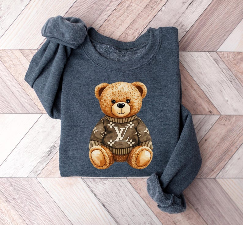 May include: A dark gray sweatshirt featuring a watercolor illustration of a teddy bear wearing a brown sweater with a white and brown logo. The bear has a friendly expression.