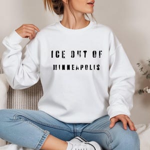 Puede incluir: Sudadera blanca con el texto negro "ICE OUT OF MINNEAPOLIS" en una fuente desgastada. La sudadera es de cuello redondo y la lleva puesta una persona. La persona lleva vaqueros azules y zapatillas blancas.