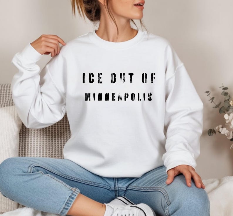 Puede incluir: Sudadera blanca con el texto negro "ICE OUT OF MINNEAPOLIS" en una fuente desgastada. La sudadera es de cuello redondo y la lleva puesta una persona. La persona lleva vaqueros azules y zapatillas blancas.