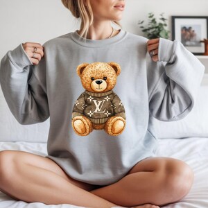 May include: A pale grey sweatshirt with a watercolour illustration of a teddy bear wearing a brown sweater with a white logo. The teddy bear has brown fur and is sitting upright.