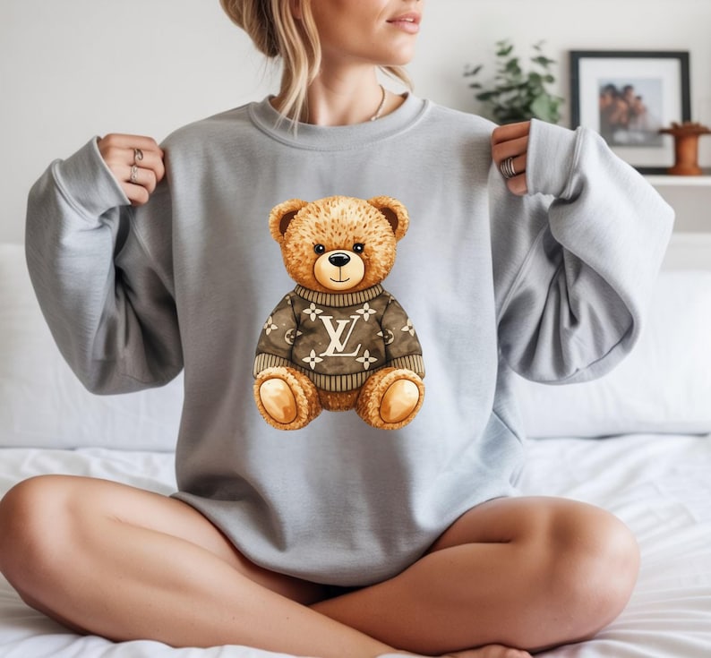 May include: A pale grey sweatshirt with a watercolour illustration of a teddy bear wearing a brown sweater with a white logo. The teddy bear has brown fur and is sitting upright.