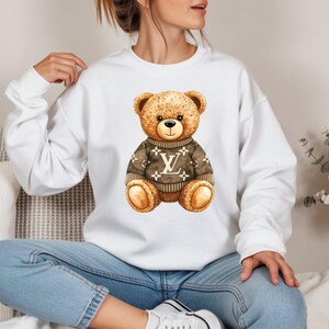 May include: A white sweatshirt with a watercolour illustration of a teddy bear wearing a brown sweater with a logo design. The bear has brown fur, a black nose, and a smiling expression. The sweatshirt is a casual style.