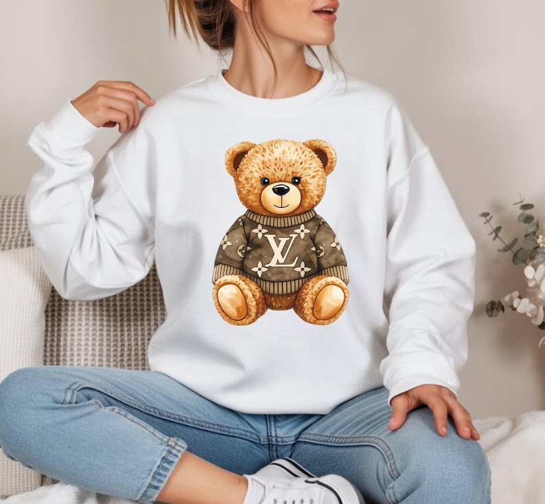 May include: A white sweatshirt with a watercolour illustration of a teddy bear wearing a brown sweater with a logo design. The bear has brown fur, a black nose, and a smiling expression. The sweatshirt is a casual style.