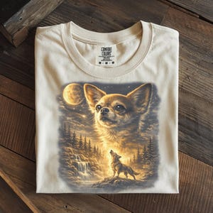 May include: Cream-colored t-shirt featuring a detailed illustration of a Chihuahua dog with a full moon and a howling wolf in a forest scene. The shirt is made of a soft material and has a Comfort Colors tag.