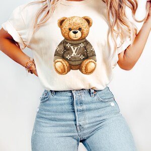 May include: Cream-coloured t-shirt featuring a brown teddy bear wearing a sweater with a logo design. The bear has a friendly expression. The shirt is paired with light blue denim jeans.