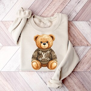 May include: Cream-coloured sweatshirt with a watercolour illustration of a teddy bear. The bear wears a brown sweater with a white logo. The bear has brown fur and a black nose, centered on the folded garment.