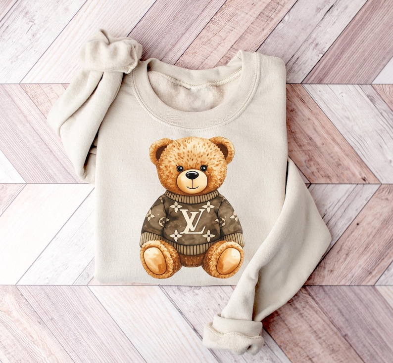 May include: Cream-coloured sweatshirt with a watercolour illustration of a teddy bear. The bear wears a brown sweater with a white logo. The bear has brown fur and a black nose, centered on the folded garment.