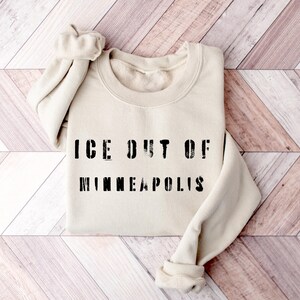 Puede incluir: Sudadera color crema con el texto negro "ICE OUT OF MINNEAPOLIS". Las mangas est&aacute;n enrolladas. El fondo es una superficie de madera de color claro.