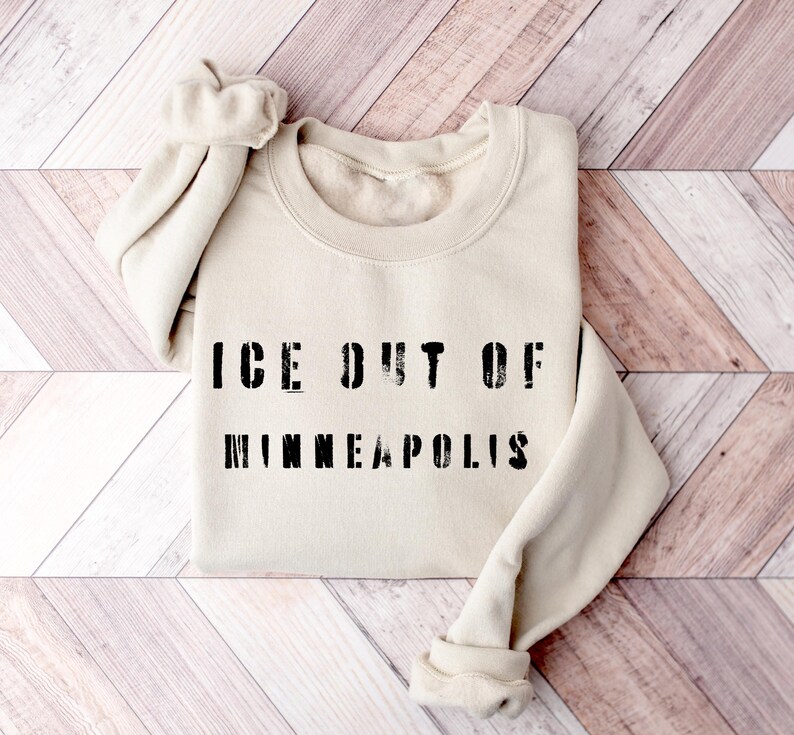 Puede incluir: Sudadera color crema con el texto negro "ICE OUT OF MINNEAPOLIS". Las mangas est&aacute;n enrolladas. El fondo es una superficie de madera de color claro.