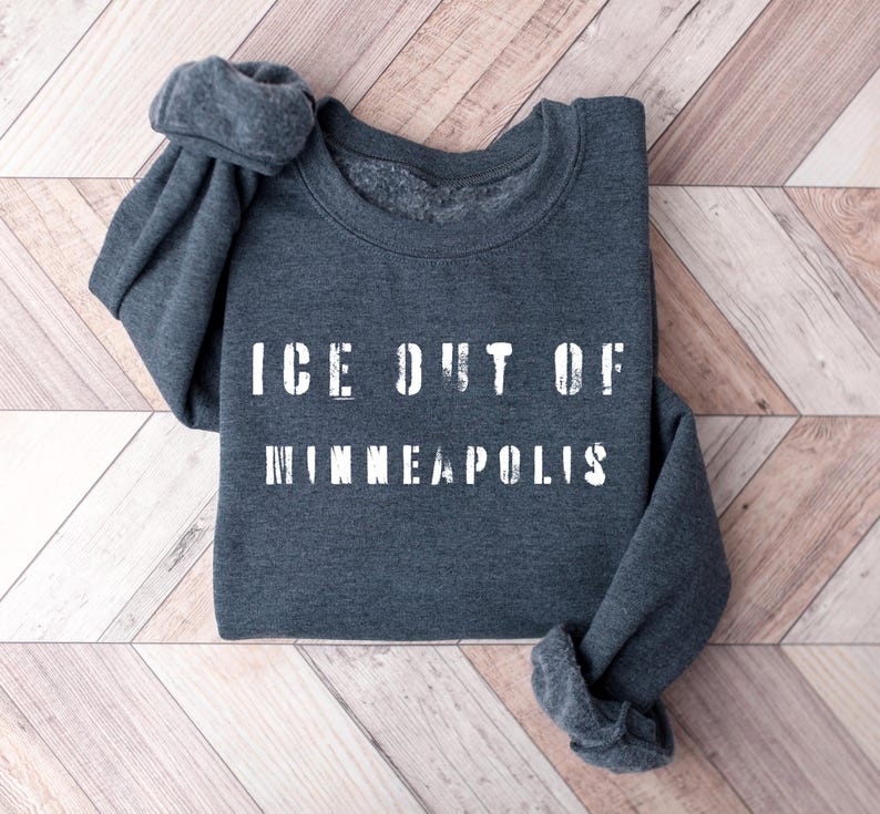 Puede incluir: Una sudadera gris oscuro con el texto blanco "ICE OUT OF MINNEAPOLIS". Las mangas est&aacute;n enrolladas. La prenda se muestra sobre una superficie de madera marr&oacute;n claro. El texto est&aacute; en una fuente de bloque.