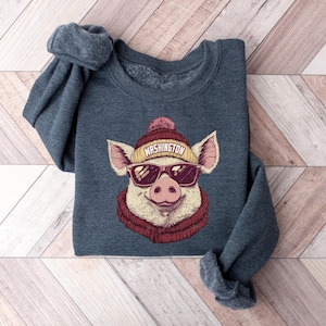 May include: A dark blue sweatshirt featuring a graphic of a pig wearing sunglasses, a knit cap with "WASHINGTON" printed on it, and a scarf. The pig is smiling. The sweatshirt is folded on a wooden surface.
