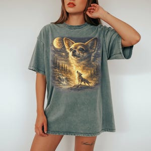 May include: A faded green t-shirt featuring a detailed graphic of a Chihuahua's face, a howling wolf, a full moon, and a waterfall. The artwork is rendered in warm, golden tones, creating a whimsical and artistic design.