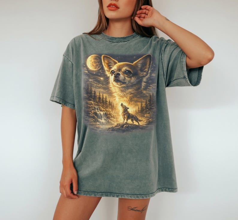 May include: A faded green t-shirt featuring a detailed graphic of a Chihuahua's face, a howling wolf, a full moon, and a waterfall. The artwork is rendered in warm, golden tones, creating a whimsical and artistic design.