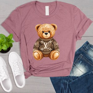 May include: Mauve t-shirt with a brown teddy bear wearing a sweater with a white logo. The bear has a black nose and is sitting. White trainers and a small green plant are visible.