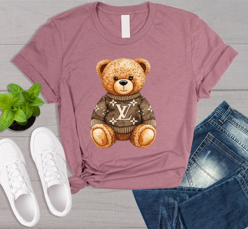 May include: Mauve t-shirt with a brown teddy bear wearing a sweater with a white logo. The bear has a black nose and is sitting. White trainers and a small green plant are visible.