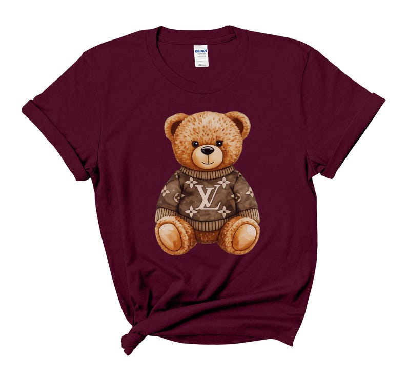 May include: A maroon t-shirt featuring a watercolor illustration of a teddy bear wearing a brown sweater with a white "LV" logo. The teddy bear has brown fur, a black nose, and is centered on the shirt.