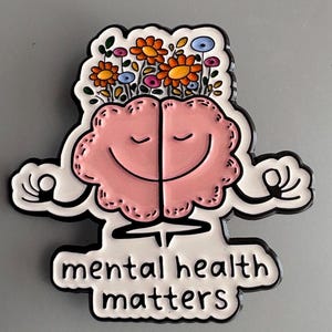 May include: A pink brain-shaped pin with a smiling face and flowers sprouting from the top. The pin has the words "mental health matters" in black text on a white background, with a black border. The pin is designed to look like a person in a meditative pose.
