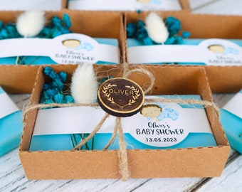 May include: A collection of baby shower favours. Each favour is a rectangular box with a light blue interior, a white label with text, and a decorative arrangement of blue and white dried flowers. The boxes are tied with twine and a wooden emblem.