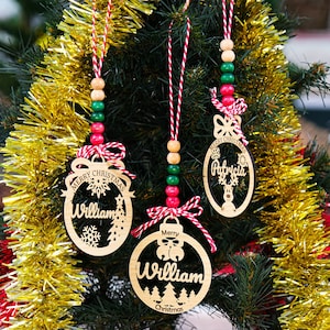 May include: Three personalized wooden Christmas ornaments with red and white twine, green and red beads, and the names "William" and "Patricia" on them. The ornaments are hanging on a Christmas tree with gold tinsel.