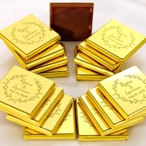 Puede incluir: Disposición de chocolates cuadrados envueltos en oro, algunos apilados, otros en abanico. Cada chocolate está grabado con "Ryan & Lauren July 14, 2025" dentro de un diseño de corona. Una caja de chocolate marrón está en el fondo.